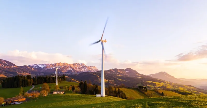 A wind turbine stands tall against a mountainous backdrop under a vibrant sky at sunset.