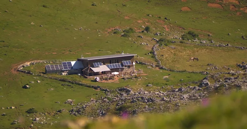 Aerial view of a sustainable cabin with solar panels, nestled in a vast, green hillside landscape.