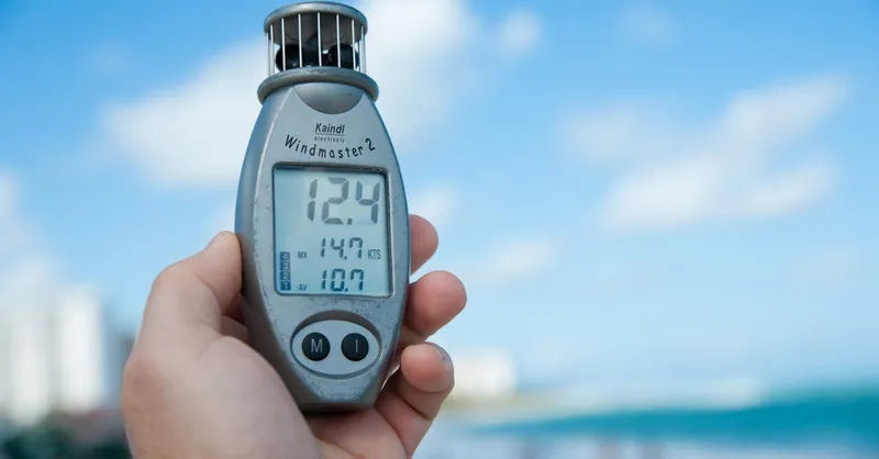 Close-up of a hand holding Windmaster 2 anemometer against a clear blue sky and beach backdrop.