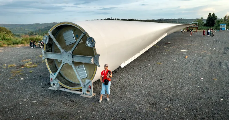 A massive wind turbine blade lying outdoors with people nearby for scale.